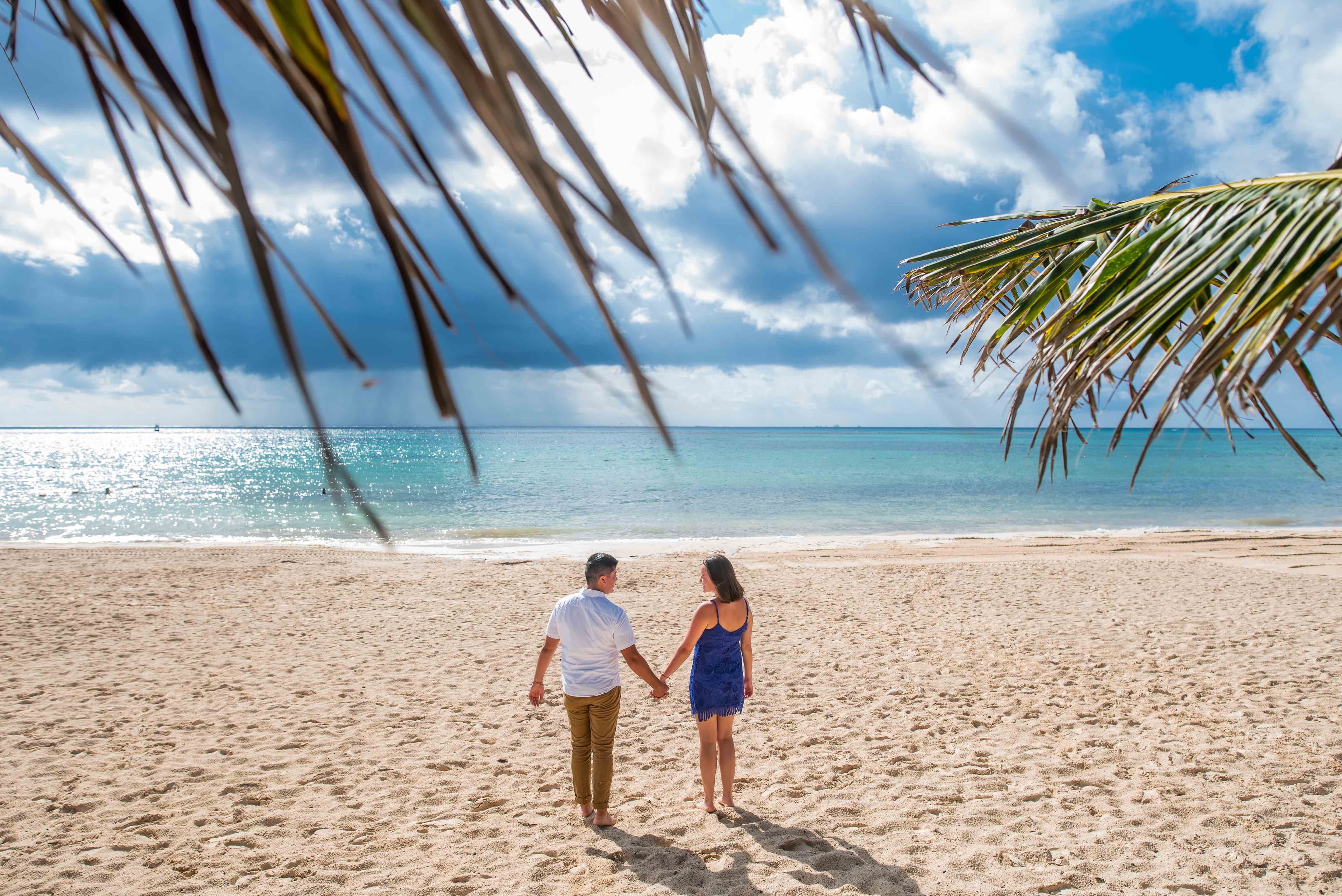 Eli and Hannah holding hands on a beach facing the ocean.