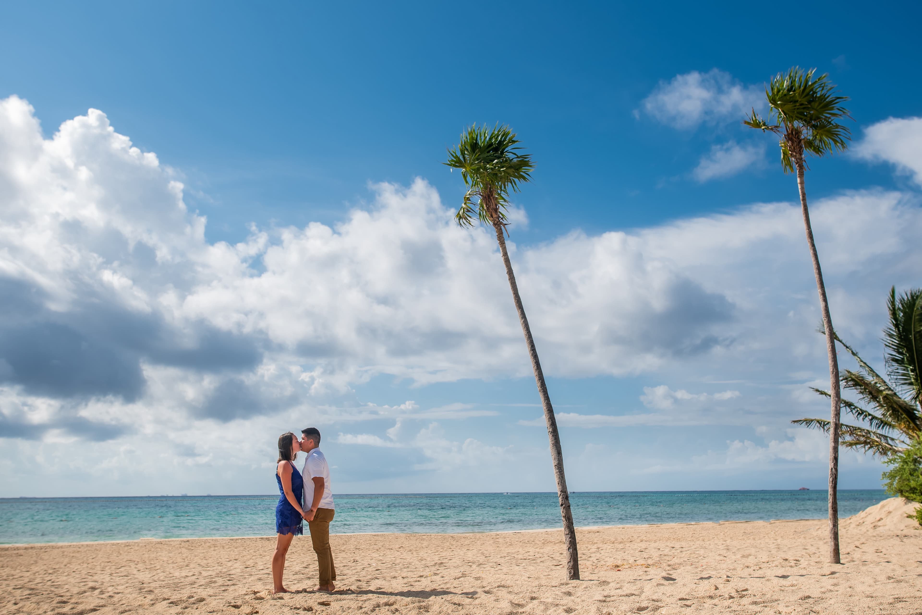 Eli and Hannah kissing on the beach near palm trees.